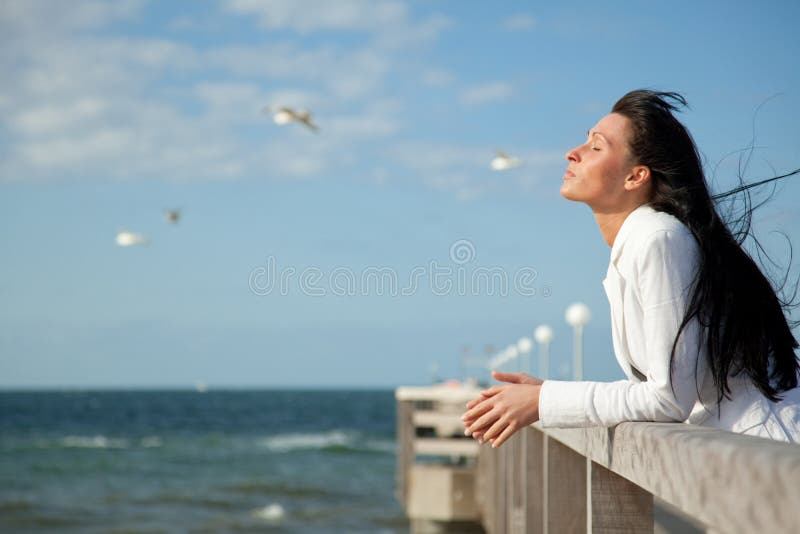 Brunette woman with flying winday hair on boardwalk looking the wide coast space with flying seagulls in background feeling free. Brunette woman with flying winday hair on boardwalk looking the wide coast space with flying seagulls in background feeling free