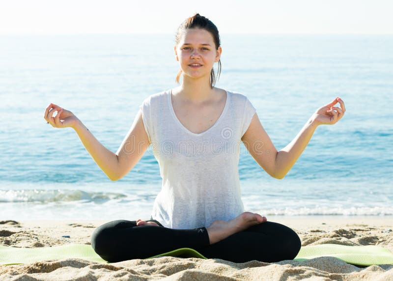 Female 20-30 years old in white T-shirt is sitting and doing meditation on the beach. Female 20-30 years old in white T-shirt is sitting and doing meditation on the beach.