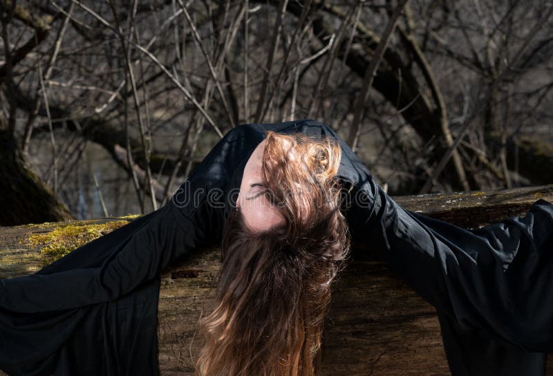 Woman with dark long hair in black robes hanging on tree branches in the middle of the forest. Back to Nature concept. Witch. Witchcraft and magic. Woman with dark long hair in black robes hanging on tree branches in the middle of the forest. Back to Nature concept. Witch. Witchcraft and magic.