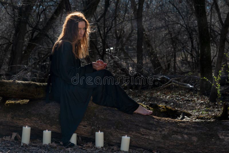 Woman with dark long hair in black robes surrounded by white burning candles sitting on a tree trunk in the forest. Back to Nature concept. Witchcraft. Woman with dark long hair in black robes surrounded by white burning candles sitting on a tree trunk in the forest. Back to Nature concept. Witchcraft.