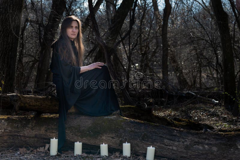 Woman with dark long hair in black robes surrounded by white burning candles sitting on a tree trunk in the forest. Back to Nature concept. Witchcraft. Woman with dark long hair in black robes surrounded by white burning candles sitting on a tree trunk in the forest. Back to Nature concept. Witchcraft.