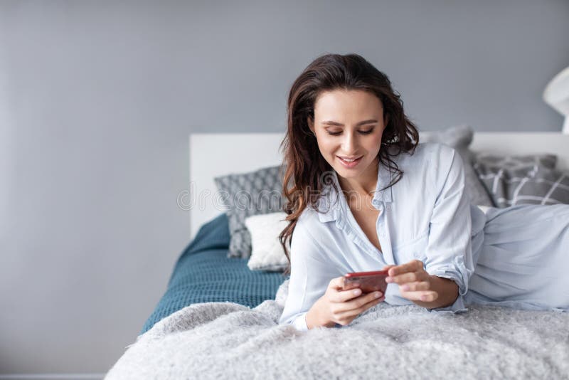 Mulher de encantamento que usa o telefone celular ao encontrar-se na cama acolhedor em casa na manhã foto de stock Mulher de encantamento que usa o telefone celular ao encontrar-se na cama acolhedor em casa na manhã foto de stock