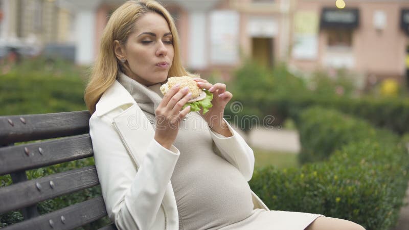 Mulher de espera bonita que come o hamburguer, banco de assento, tocando na barriga, apetite