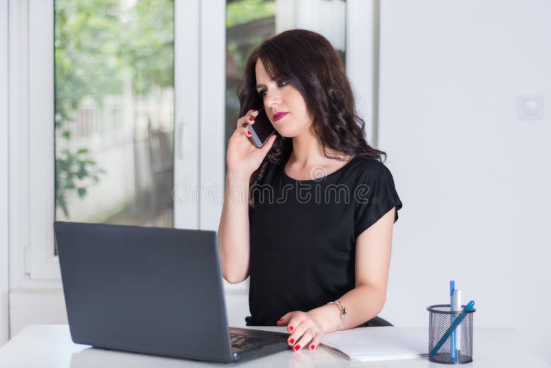 Mulher de negócio bonito no escritório que fala no telefone celular e que trabalha no portátil foto de stock Mulher de negócio bonito no escritório que fala no telefone celular e que trabalha no portátil foto de stock