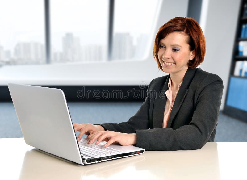Mulher de negócio feliz com cabelo vermelho que sorri no trabalho que datilografa no portátil do computador na mesa de escritório fotos de stock Mulher de negócio feliz com cabelo vermelho que sorri no trabalho que datilografa no portátil do computador na mesa de escritório fotos de stock