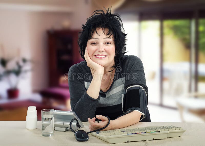 Portrait of cheerful mature woman measures her blood pressure. Smiling black haired woman sits at white desk and looks at the camera. There are pills plastic bottle, keyboard, glass water and blood pressure gauge on the desk. Horizontal shot on blurry indoors background. Portrait of cheerful mature woman measures her blood pressure. Smiling black haired woman sits at white desk and looks at the camera. There are pills plastic bottle, keyboard, glass water and blood pressure gauge on the desk. Horizontal shot on blurry indoors background