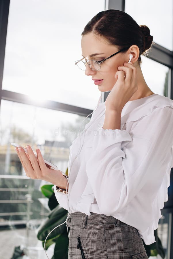 Mulher de negócios nova que está na frente da janela, ao escutar a música nos earbuds imagem de stock royalty free Mulher de negócios nova que está na frente da janela, ao escutar a música nos earbuds imagem de stock royalty free