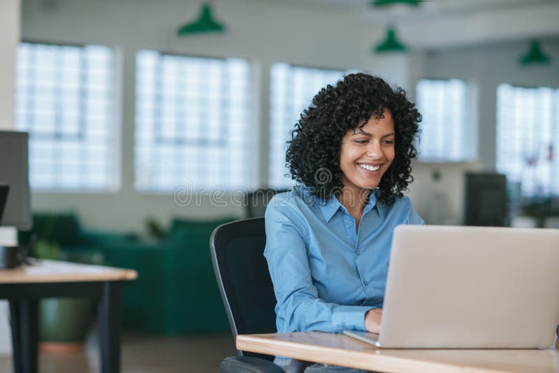 Mulher de negócios nova de sorriso que trabalha em um portátil em sua mesa imagem de stock royalty free Mulher de negócios nova de sorriso que trabalha em um portátil em sua mesa imagem de stock royalty free