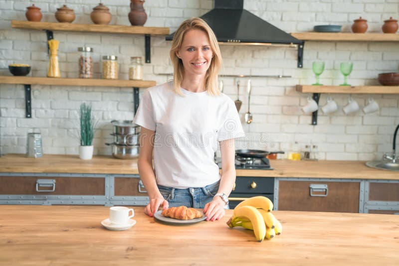 A mulher de sorriso nova bonita está preparando o café da manhã na cozinha em casa café da manhã, croissant, bananas Alimento sau fotografia de stock A mulher de sorriso nova bonita está preparando o café da manhã na cozinha em casa café da manhã, croissant, bananas Alimento sau fotografia de stock