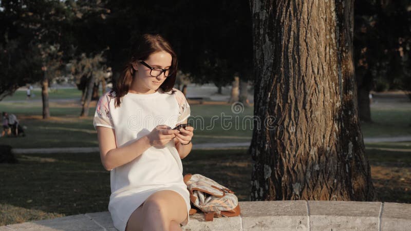Mulher de sorriso nova que usa o smartphone que senta-se no banco no parque no por do sol Menina europeia bonita que texting no t imagem de stock royalty free Mulher de sorriso nova que usa o smartphone que senta-se no banco no parque no por do sol Menina europeia bonita que texting no t imagem de stock royalty free
