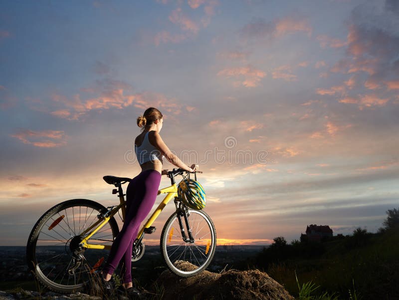 A mulher delgada com Mountain bike está estando no monte sob o céu bonito no por do sol fotos de stock royalty free A mulher delgada com Mountain bike está estando no monte sob o céu bonito no por do sol fotos de stock royalty free