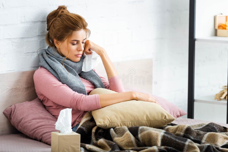 Mulher doente com cachecol cinza sentada na cama e segurando guardanapo fotografia de stock royalty free