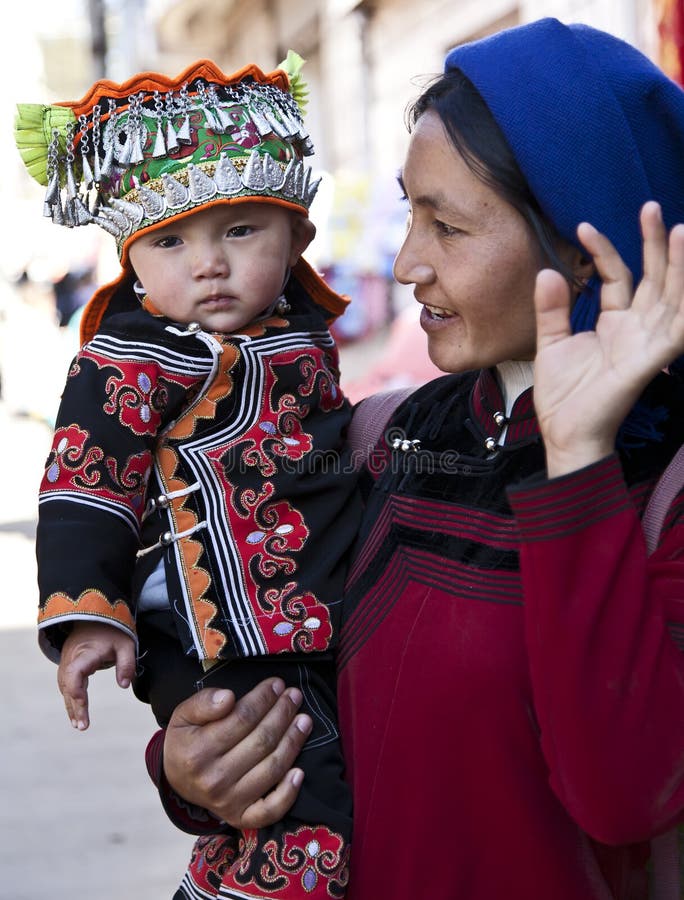 A baby boy wearing the traditional, elaborate hats of the Hani people as his mother waves to the camera at a market in southern Yunnan province in China. A baby boy wearing the traditional, elaborate hats of the Hani people as his mother waves to the camera at a market in southern Yunnan province in China.