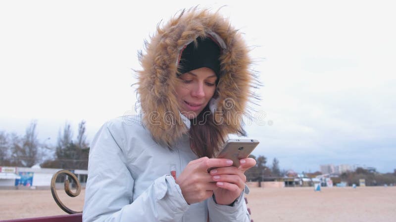 Mulher em um revestimento branco da pena que senta-se em um banco na praia e que datilografa uma mensagem no telefone celular