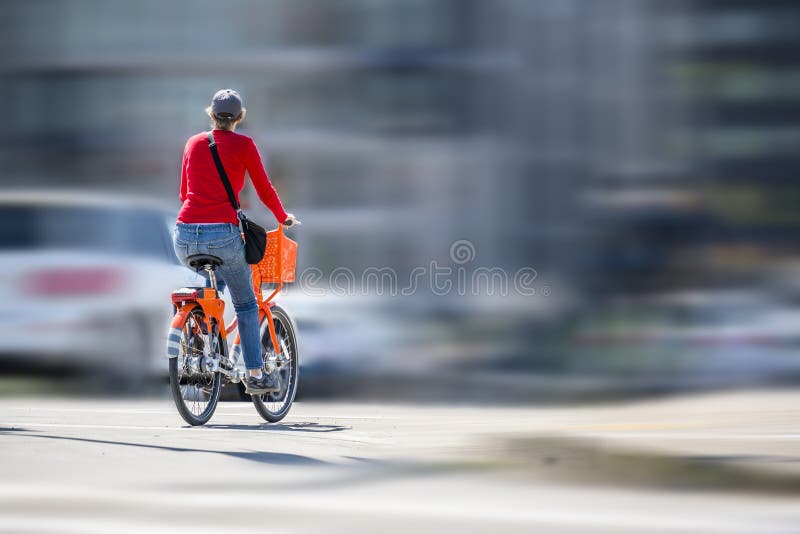 Mulher em uma bicicleta alaranjada com passeios da cesta no trajeto da bicicleta na estrada ao lado de outros veículos fotografia de stock Mulher em uma bicicleta alaranjada com passeios da cesta no trajeto da bicicleta na estrada ao lado de outros veículos fotografia de stock