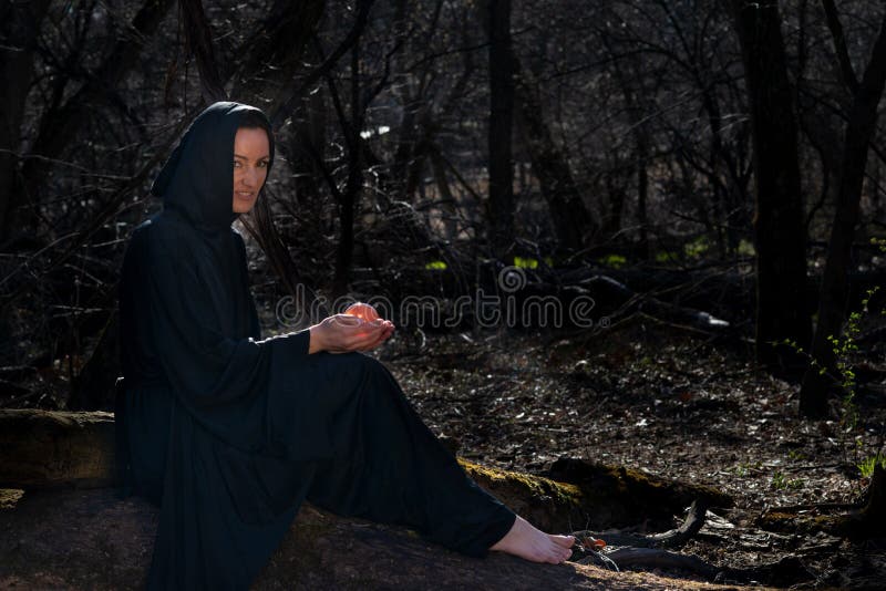 Hooded Woman with dark long hair in black robes sitting on a tree trunk in the forest. Back to Nature concept. Witchcraft and magic. Scary scenery. Hooded Woman with dark long hair in black robes sitting on a tree trunk in the forest. Back to Nature concept. Witchcraft and magic. Scary scenery.