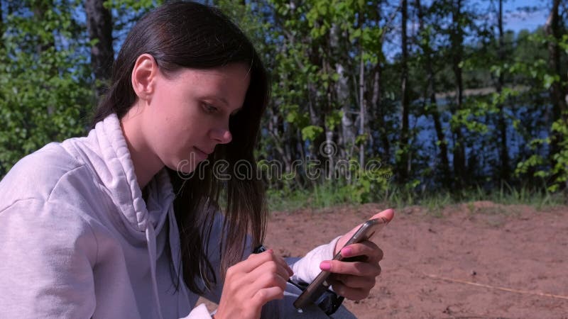 A mulher está digitando mensagens no smartphone sentado na natureza, vista lateral