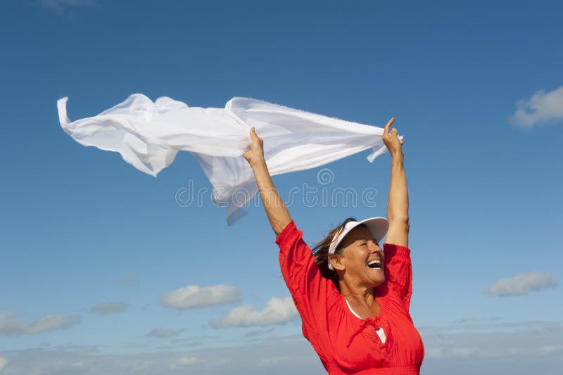An attractive looking mature woman cheerful and happy laughing while holding a light white fluttering fabric in the wind, with twilight sunshine and blue sky as background and copy space. An attractive looking mature woman cheerful and happy laughing while holding a light white fluttering fabric in the wind, with twilight sunshine and blue sky as background and copy space.