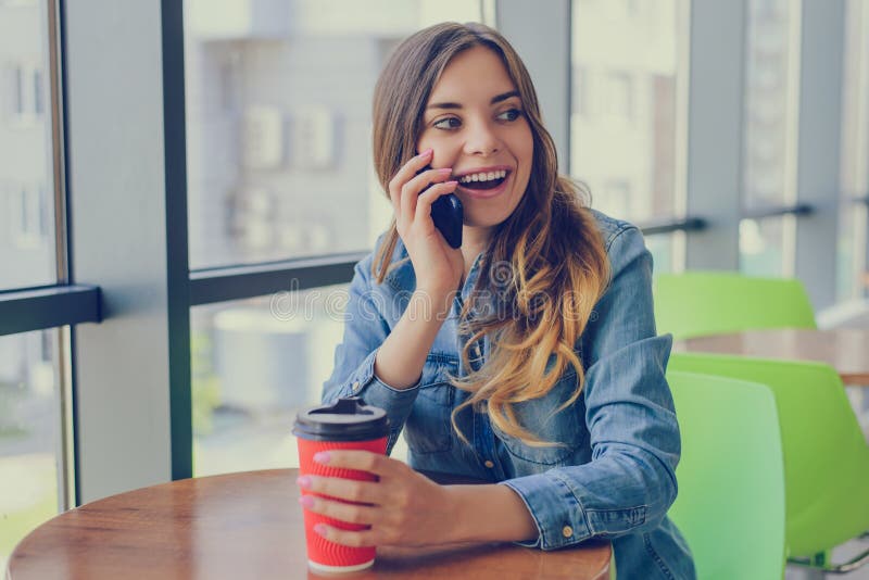 Mulher feliz de sorriso entusiástica que senta-se em um café que guarda a xícara de café, está falando no telefone com amigo, con foto de stock royalty free Mulher feliz de sorriso entusiástica que senta-se em um café que guarda a xícara de café, está falando no telefone com amigo, con foto de stock royalty free