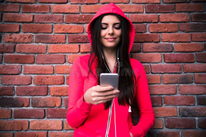 Mulher feliz que usa o smartphone sobre a parede de tijolo