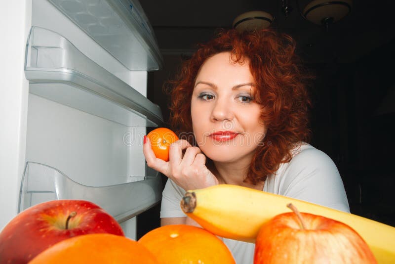 A mulher grande come o fruto Menina gorda do cabelo vermelho que olha o refrigerat interno