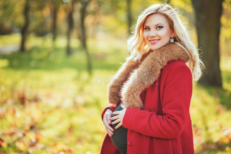 Mulher gravida feliz bonita que fica no parque do outono que toca em sua barriga e que aprecia a espera do bebê fotografia de stock royalty free Mulher gravida feliz bonita que fica no parque do outono que toca em sua barriga e que aprecia a espera do bebê fotografia de stock royalty free