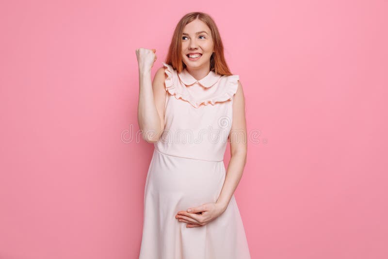 A mulher gravida feliz exulta e guarda a barriga, mostrando o gesto do sucesso e a vitória em um fundo cor-de-rosa fotos de stock royalty free A mulher gravida feliz exulta e guarda a barriga, mostrando o gesto do sucesso e a vitória em um fundo cor-de-rosa fotos de stock royalty free