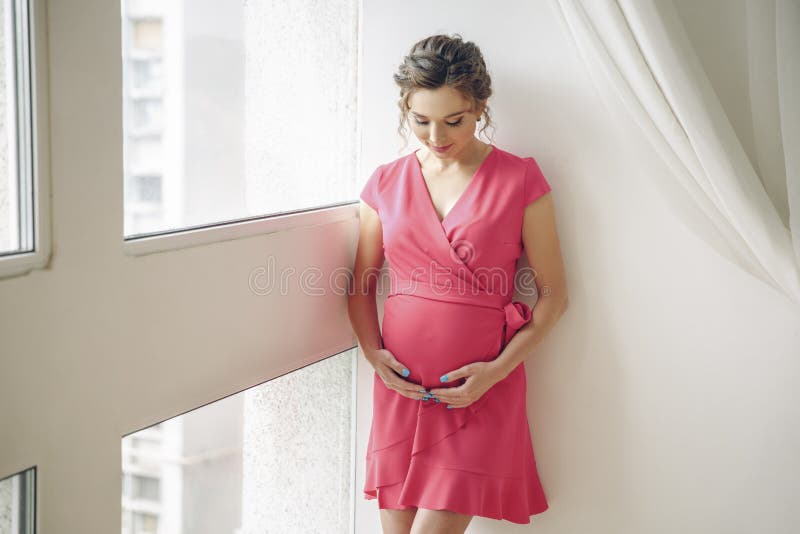 Mulher gravida nova que fica perto da janela no vestido cor-de-rosa fotos de stock Mulher gravida nova que fica perto da janela no vestido cor-de-rosa fotos de stock