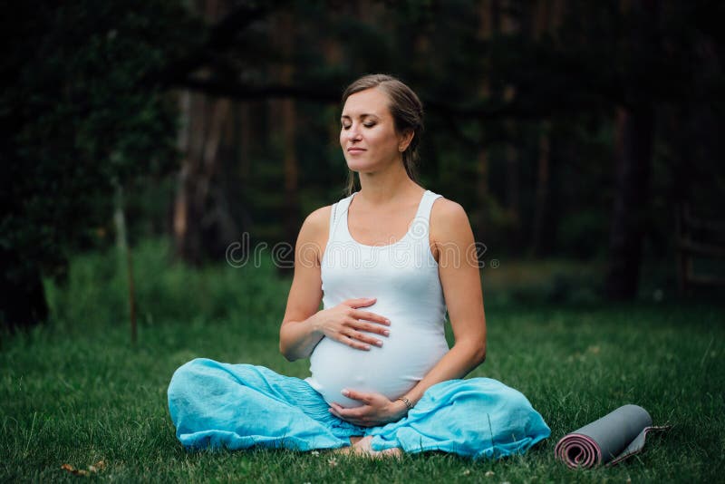 Mulher grávida da ioga com o retrato da esteira no parque na grama, respiração, esticando, estática exterior, floresta foto de stock Mulher grávida da ioga com o retrato da esteira no parque na grama, respiração, esticando, estática exterior, floresta foto de stock