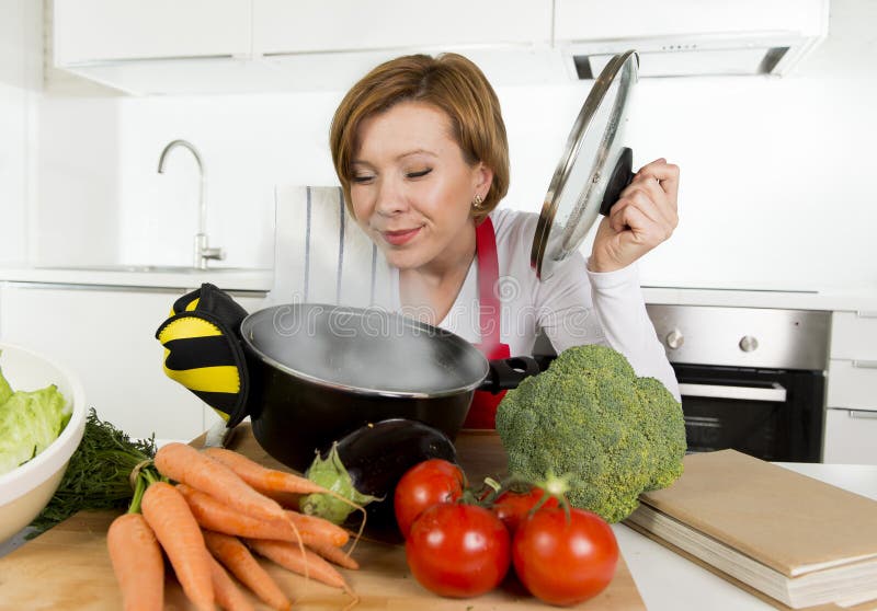 Mulher home do cozinheiro no avental vermelho na cozinha doméstica que guarda o cozimento do potenciômetro com o guisado vegetal