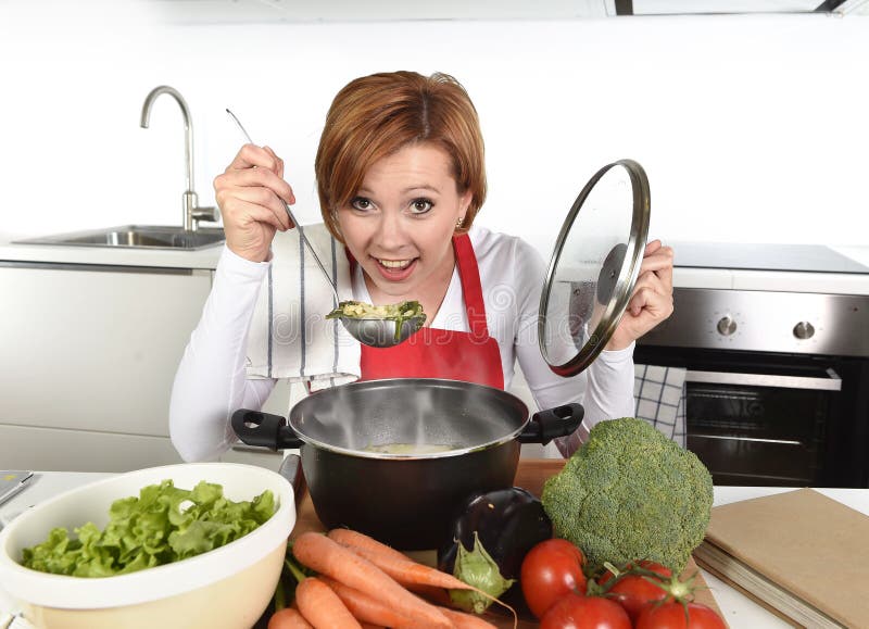 Mulher home feliz do cozinheiro no avental vermelho na cozinha doméstica que guarda a caçarola com a sopa que prova o guisado veg foto de stock royalty free Mulher home feliz do cozinheiro no avental vermelho na cozinha doméstica que guarda a caçarola com a sopa que prova o guisado veg foto de stock royalty free