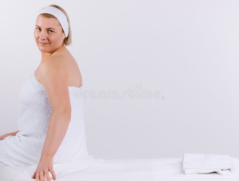 Senior woman, wrapped in a white towel, sits on a massage table before the procedure and looks cute at the camera. White background with empty space for advertising. High quality photo. Senior woman, wrapped in a white towel, sits on a massage table before the procedure and looks cute at the camera. White background with empty space for advertising. High quality photo