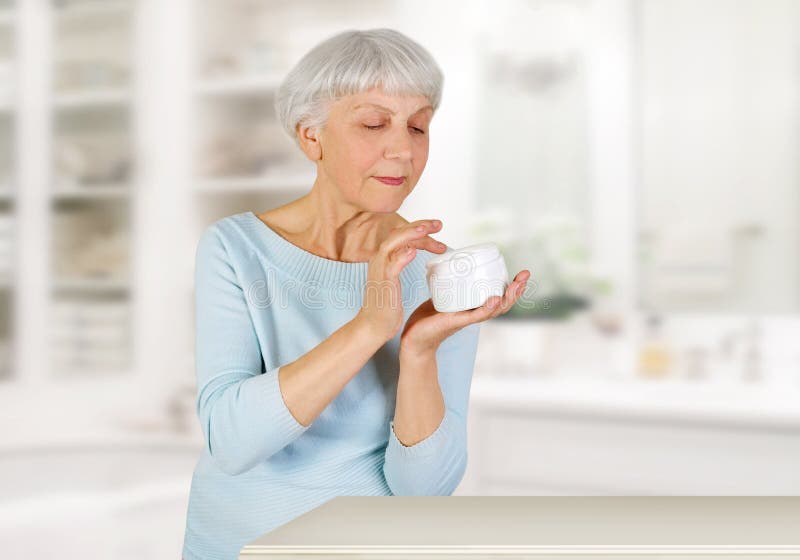 Mulher idosa encantador que aplica o creme cosmético em sua cara para cuidados com a pele faciais no banheiro em casa fotografia de stock Mulher idosa encantador que aplica o creme cosmético em sua cara para cuidados com a pele faciais no banheiro em casa fotografia de stock