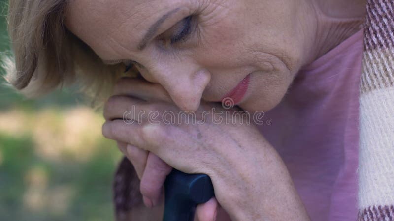 Mulher idosa triste que senta-se no parque com vara de passeio, sentindo só e infeliz