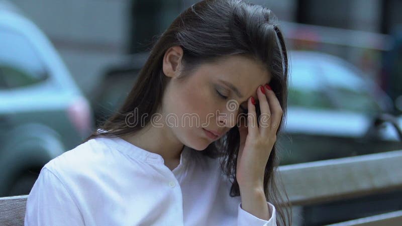 Mulher jovem sentindo dor de cabeça sentada no banco, sintoma de enxaqueca, saúde no estresse