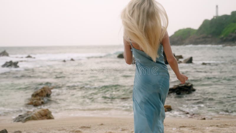 Mulher loira em vestidos azuis caminha em ondas de praia arenosas que caem na orla. pov segue viajante explorando a costa tropical