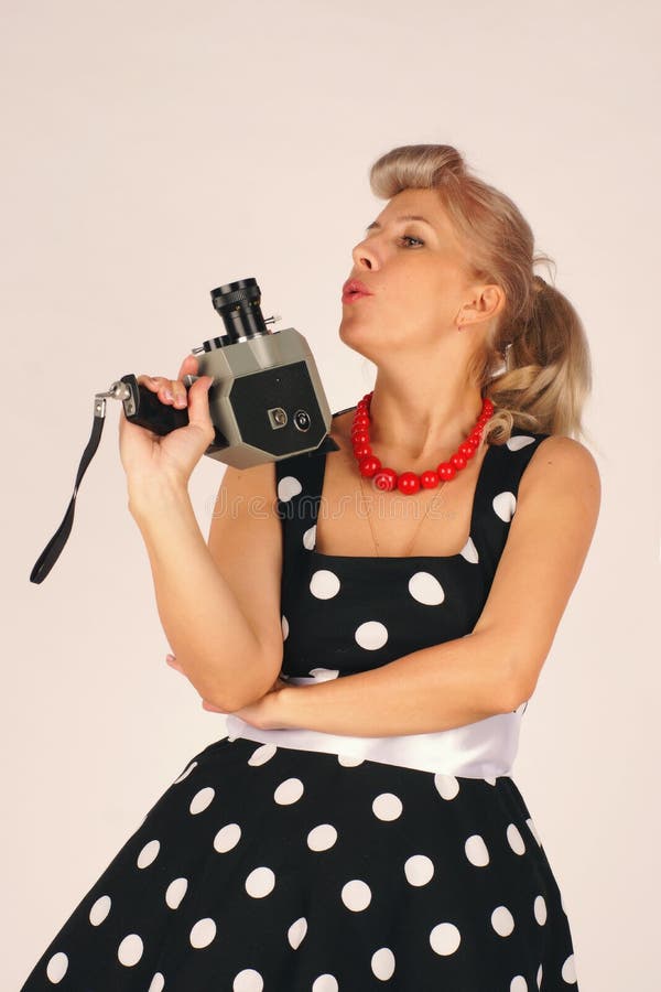 A mulher loura bonita no estilo do pinup, vestido em um vestido do às bolinhas, está e guarda uma câmera retro com uma mão com a imagem de stock royalty free A mulher loura bonita no estilo do pinup, vestido em um vestido do às bolinhas, está e guarda uma câmera retro com uma mão com a imagem de stock royalty free