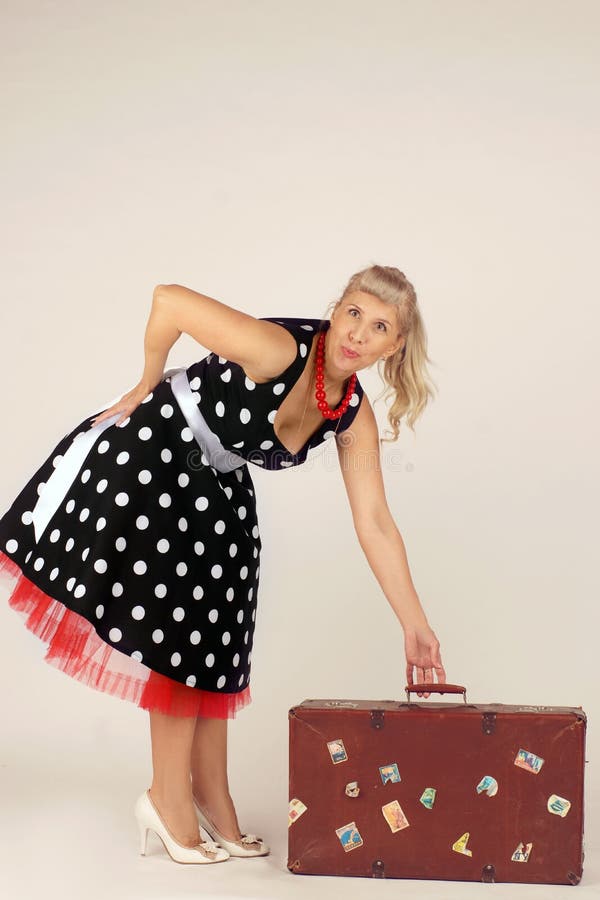 A mulher loura bonita no estilo do pinup, vestido em um vestido do às bolinhas, está e levanta uma mala de viagem do curso, fundo imagem de stock royalty free A mulher loura bonita no estilo do pinup, vestido em um vestido do às bolinhas, está e levanta uma mala de viagem do curso, fundo imagem de stock royalty free