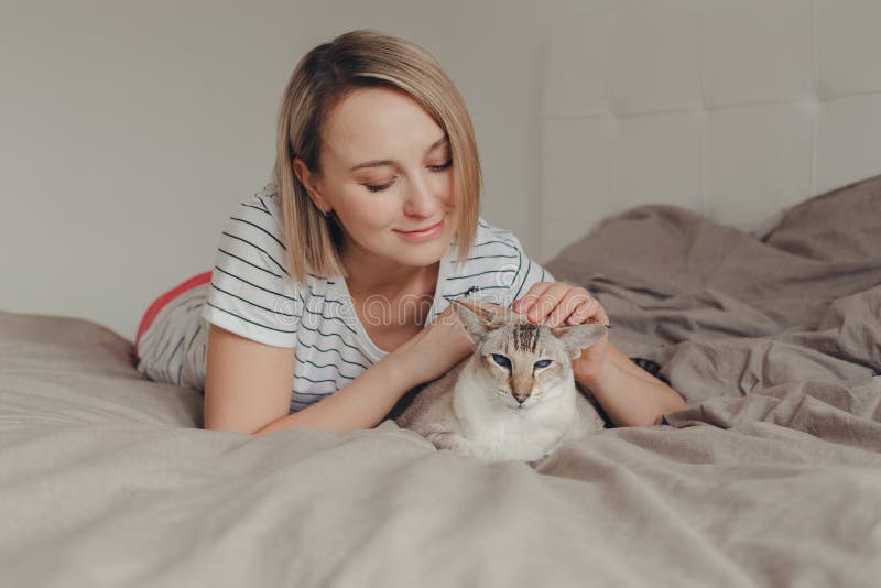 Mulher loura de sorriso caucasiano que encontram-se na cama no quarto em casa e trocas de carícias que afagam o gato ponto-colori foto de stock royalty free Mulher loura de sorriso caucasiano que encontram-se na cama no quarto em casa e trocas de carícias que afagam o gato ponto-colori foto de stock royalty free