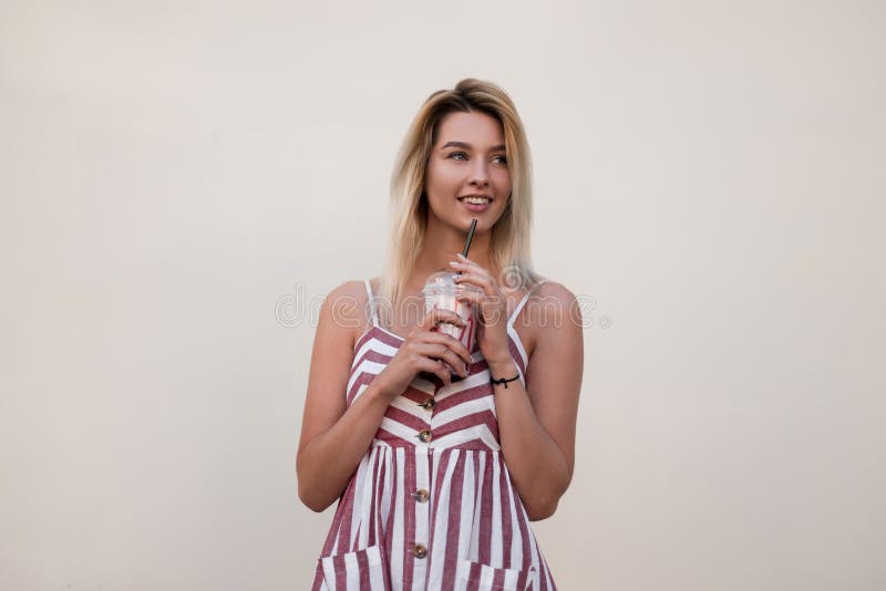 Happy pretty beautiful young blond woman in a stylish summer pink striped dress with a sweet cold milk drink rest near a vintage wall in the city. Joyful cute girl model enjoys outdoors travels. Happy pretty beautiful young blond woman in a stylish summer pink striped dress with a sweet cold milk drink rest near a vintage wall in the city. Joyful cute girl model enjoys outdoors travels