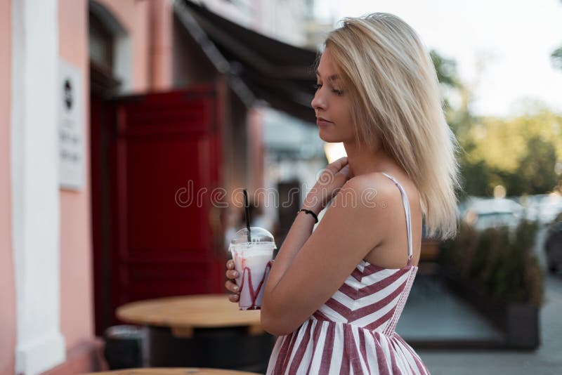 Mulher loura nova bonita moderna bonita em um vestido listrado cor-de-rosa à moda com uma caminhada fria doce da bebida de leite foto de stock Mulher loura nova bonita moderna bonita em um vestido listrado cor-de-rosa à moda com uma caminhada fria doce da bebida de leite foto de stock