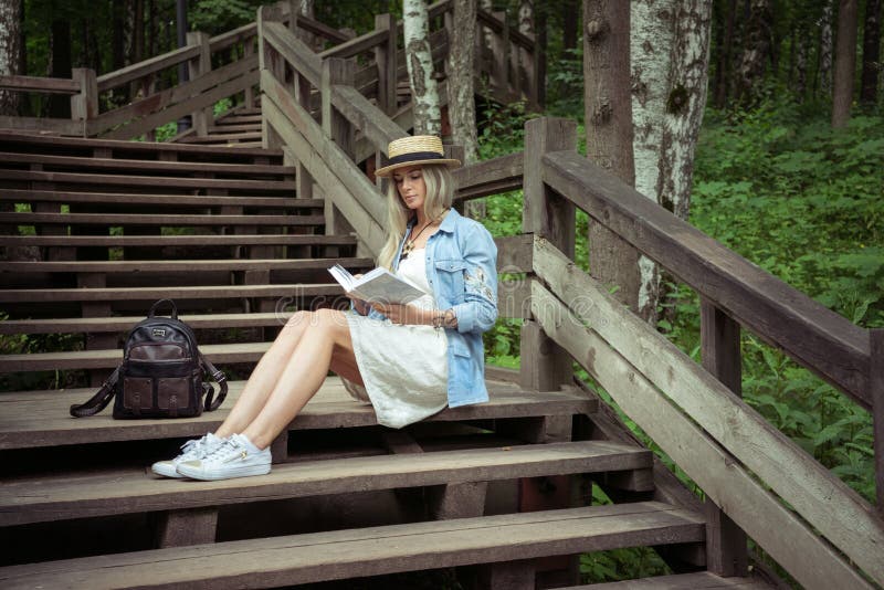 Mulher loura nova bonita que senta-se em escadas de madeira no parque da cidade e que lê um livro Tem um vestido branco, uma palh fotos de stock Mulher loura nova bonita que senta-se em escadas de madeira no parque da cidade e que lê um livro Tem um vestido branco, uma palh fotos de stock