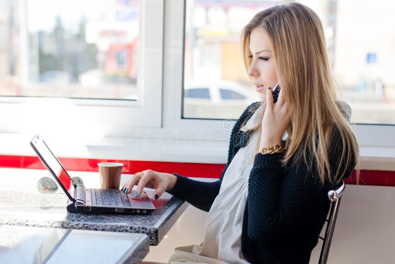 Mulher loura nova bonita séria da mulher de negócio que fala no telefone celular móvel que trabalha em um computador do PC do por imagens de stock royalty free Mulher loura nova bonita séria da mulher de negócio que fala no telefone celular móvel que trabalha em um computador do PC do por imagens de stock royalty free
