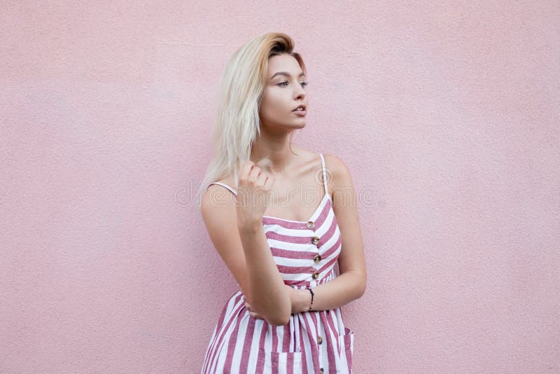 A mulher loura nova bonita urbana em sundress listrados à moda de um verão descansa perto de uma parede cor-de-rosa do vintage na imagem de stock royalty free A mulher loura nova bonita urbana em sundress listrados à moda de um verão descansa perto de uma parede cor-de-rosa do vintage na imagem de stock royalty free