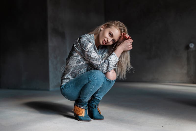 Sensual sexy young blonde woman in vintage cowboy boots in summer shirt in fashionable ripped jeans sit in a room with sunlight. American beautiful fashion model girl enjoys the rest. Sensual sexy young blonde woman in vintage cowboy boots in summer shirt in fashionable ripped jeans sit in a room with sunlight. American beautiful fashion model girl enjoys the rest