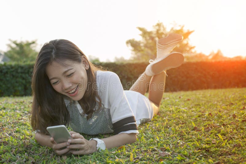Mulher mais nova asiática que encontra-se no campo que joga o telefone esperto e o riso fotografia de stock Mulher mais nova asiática que encontra-se no campo que joga o telefone esperto e o riso fotografia de stock