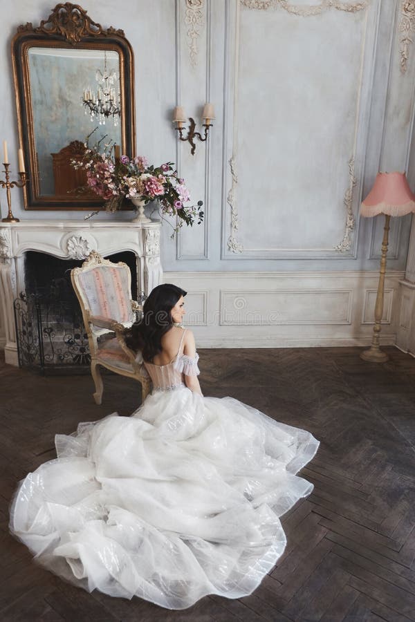 A mulher modelo moreno nova em um vestido de casamento à moda do laço com ombros despidos senta-se no assoalho perto da chaminé d imagens de stock A mulher modelo moreno nova em um vestido de casamento à moda do laço com ombros despidos senta-se no assoalho perto da chaminé d imagens de stock