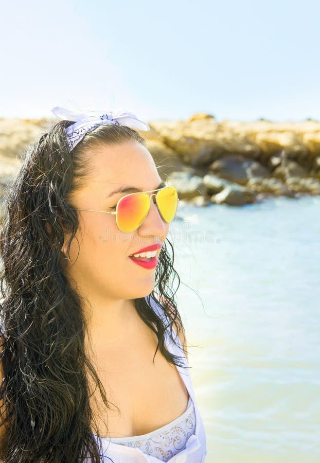 A mulher moreno nova bonita com cabelo encaracolado longo no vestido branco dos óculos de sol coloridos do espelho está no riso d fotografia de stock A mulher moreno nova bonita com cabelo encaracolado longo no vestido branco dos óculos de sol coloridos do espelho está no riso d fotografia de stock