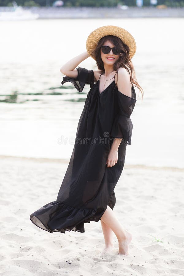 Mulher moreno nova bonita no vestido preto, em óculos de sol pretos e em chapéu de palha andando na areia na praia e foto de stock Mulher moreno nova bonita no vestido preto, em óculos de sol pretos e em chapéu de palha andando na areia na praia e foto de stock