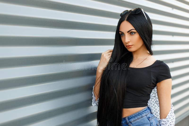 Mulher moreno nova moderna bonita bonita com cabelo longo em calças de brim do vintage em uma parte superior preta à moda em um d imagens de stock Mulher moreno nova moderna bonita bonita com cabelo longo em calças de brim do vintage em uma parte superior preta à moda em um d imagens de stock